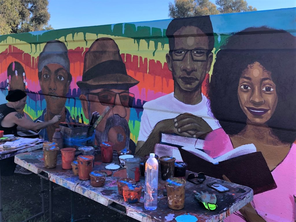 Portion of Teralta Park Unity in Community Mural featuring images of people from the City Heights community; paint supplies sit in front of the mural