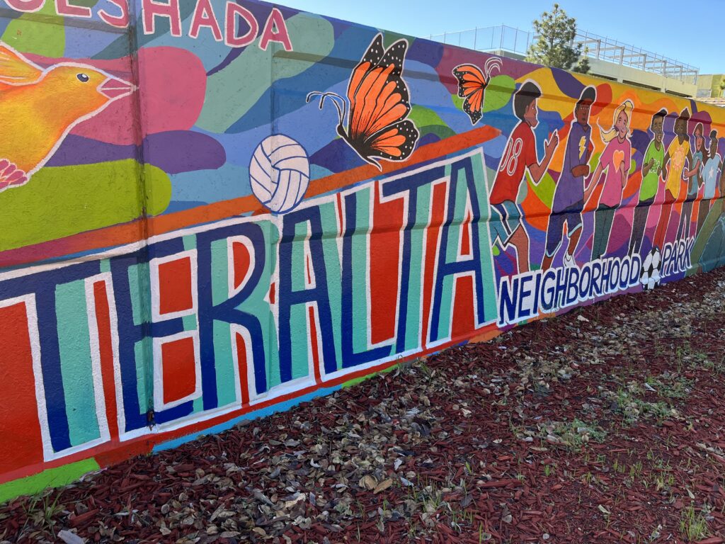 Portion of Teralta Park Unity mural featuring the words "Teralta Neighborhood Park", a volleyball, a butterfly, and a group of children in multicolored clothes running and playing.