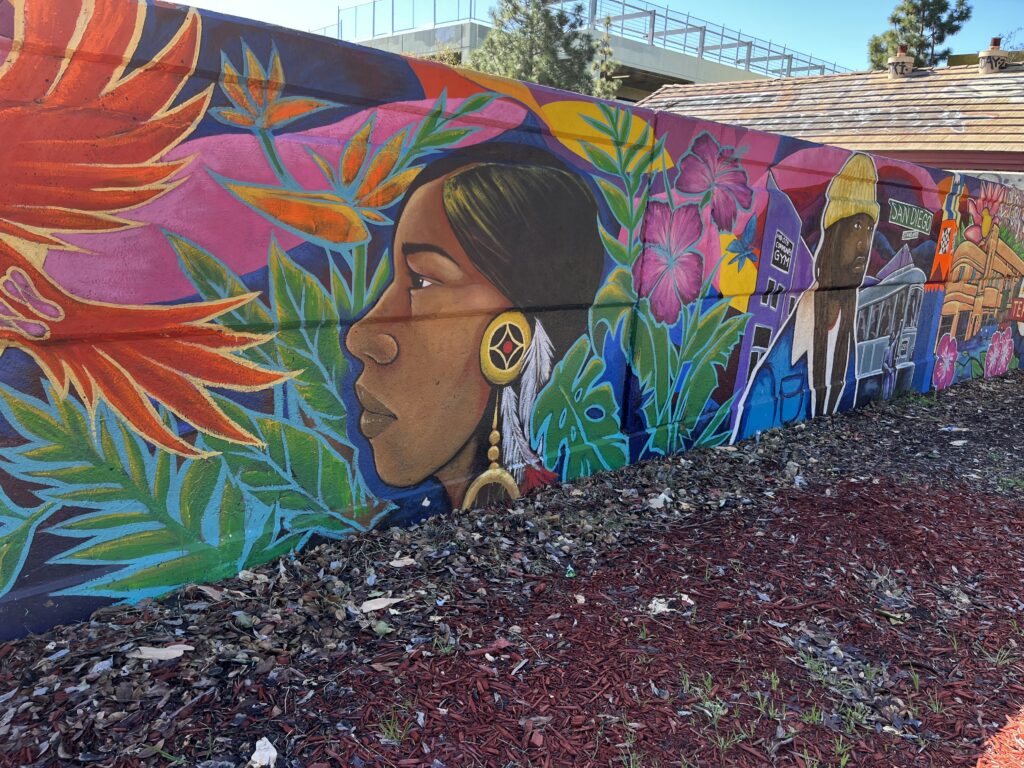Portion of Teralta Park Unity mural featuring profile of a Native American person, vibrant plants and flowers, and an black man wearing a blue jacket and a yellow hat.