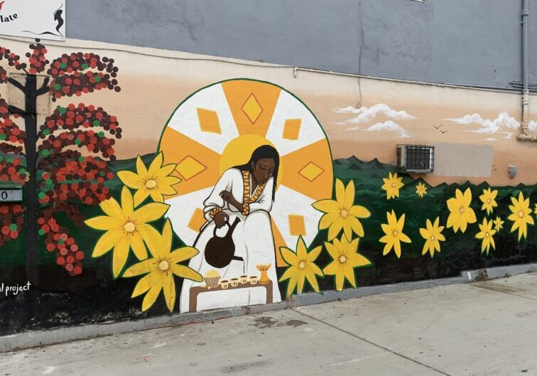 Colorful urban wall mural depicting a woman pouring coffee, surrounded by vibrant yellow flowers.